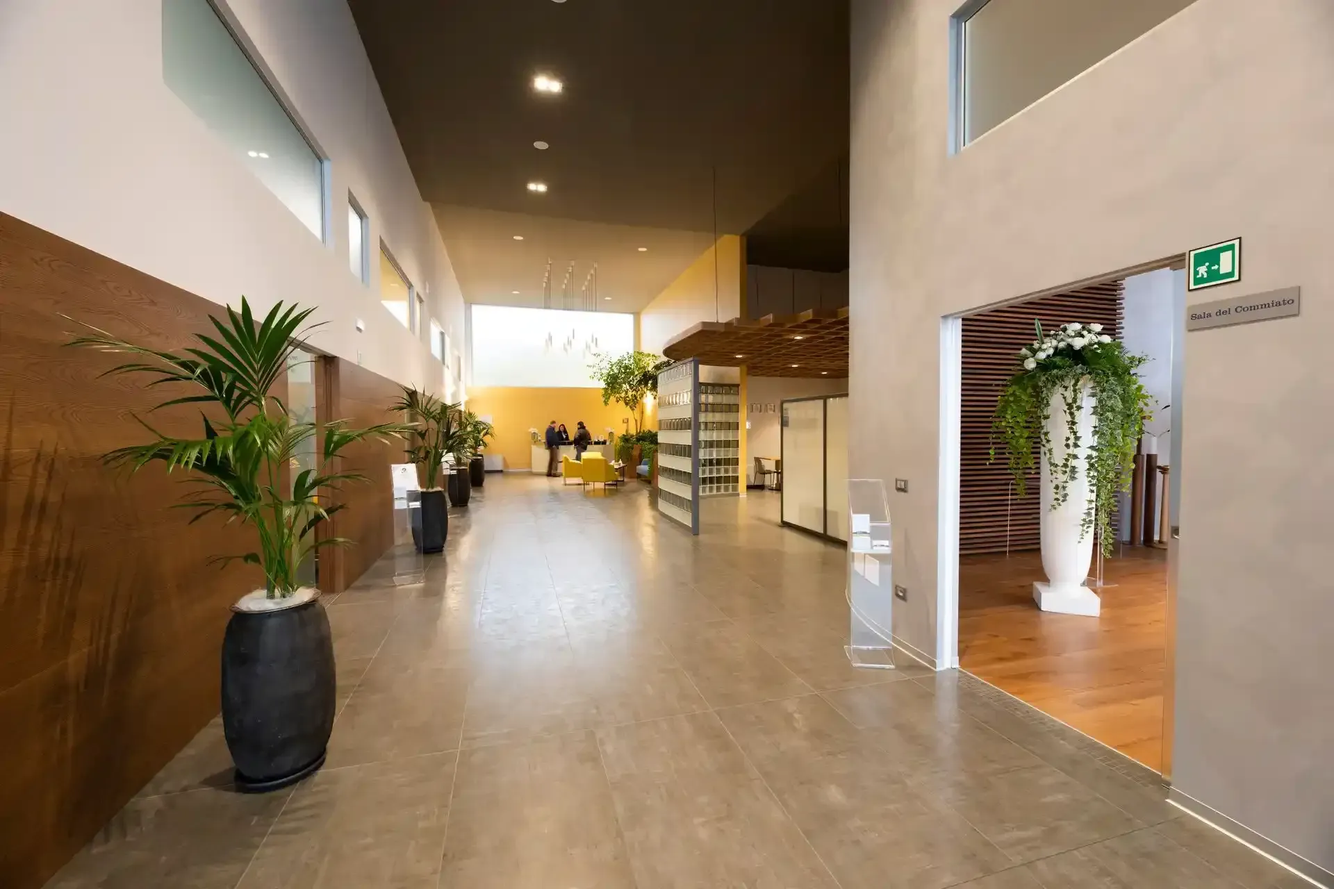 Lungo corridoio con piante in vaso, verso una zona con pareti in vetro. Ingresso sulla destra, con esposizione floreale.