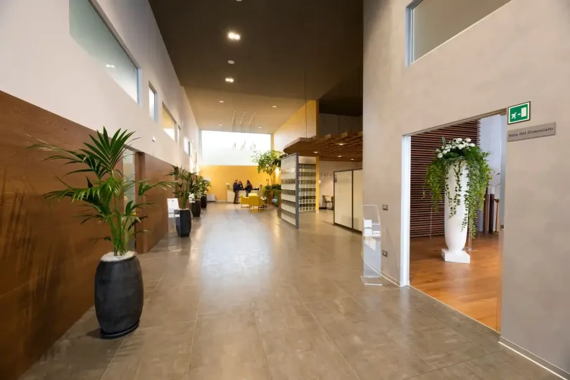 Lungo corridoio con piante in vaso, verso una zona con pareti in vetro. Ingresso sulla destra, con esposizione floreale.