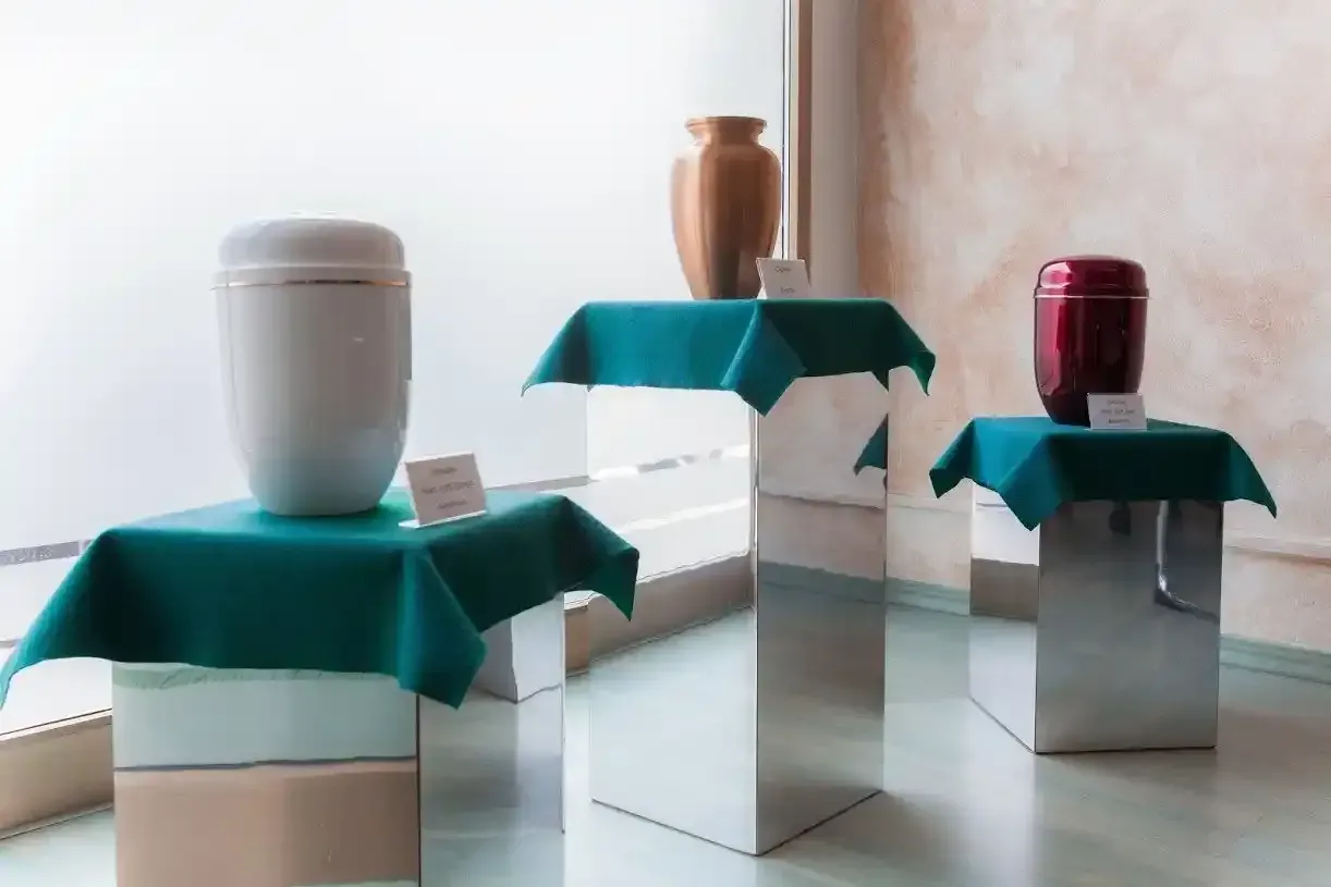 Tre urne cinerarie su piedistalli. Urne bianche, bronzo e rosse sono appoggiate su teli verdi, sopra supporti metallici.