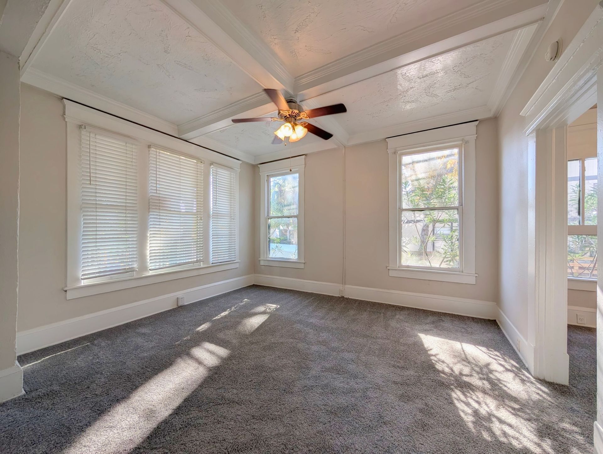 Empty room with gray carpet, white trim, windows, ceiling fan, and sunlight.