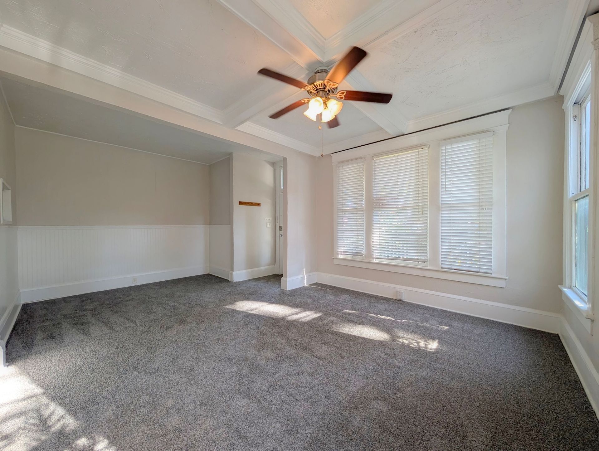 Empty room with gray carpet, white walls, and a ceiling fan. Natural light streams through the window.