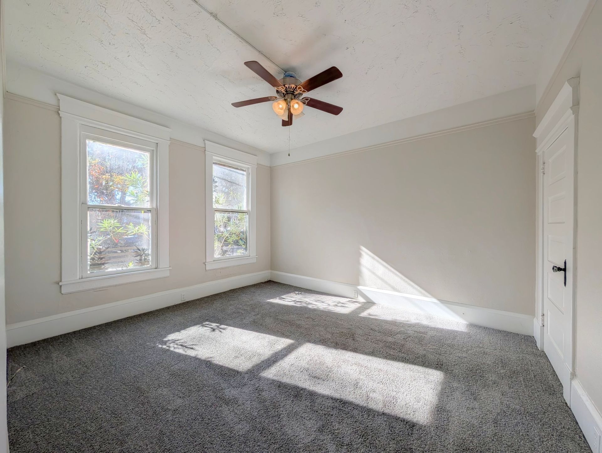 Empty bedroom with windows, ceiling fan, and gray carpet. Sunlight shines on the floor.