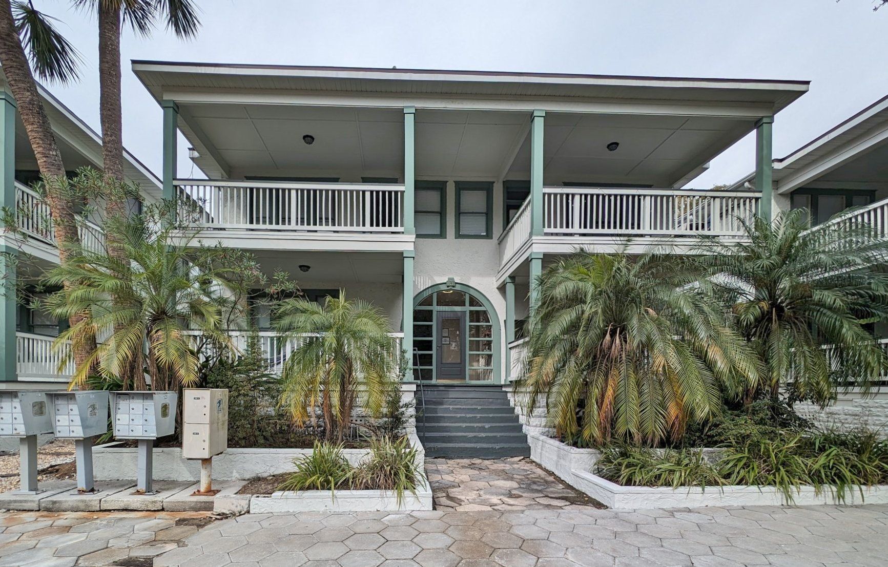 Two-story building with balconies, green accents, and palm trees in front.
