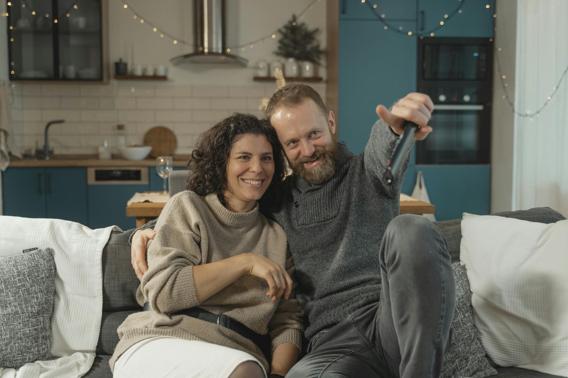 Couple smiling, sitting on a couch, man holding remote. Kitchen background.