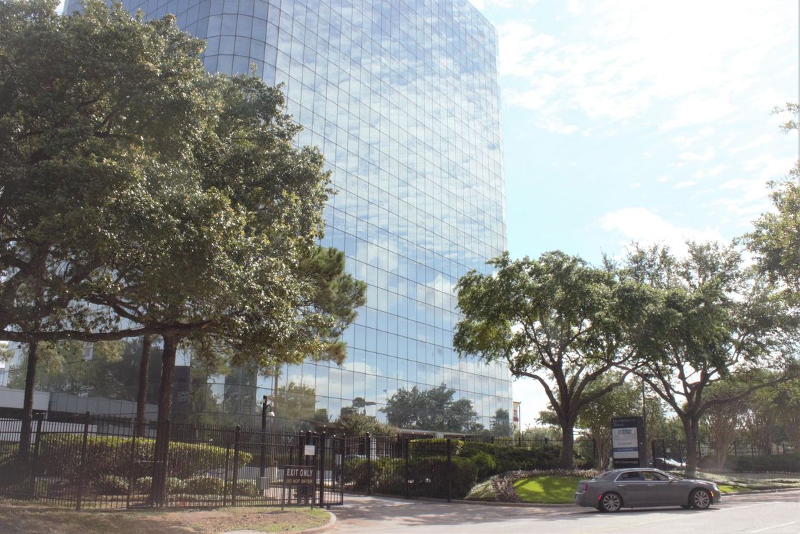 Glass skyscraper with trees in front, partly cloudy sky, car parked in lot.