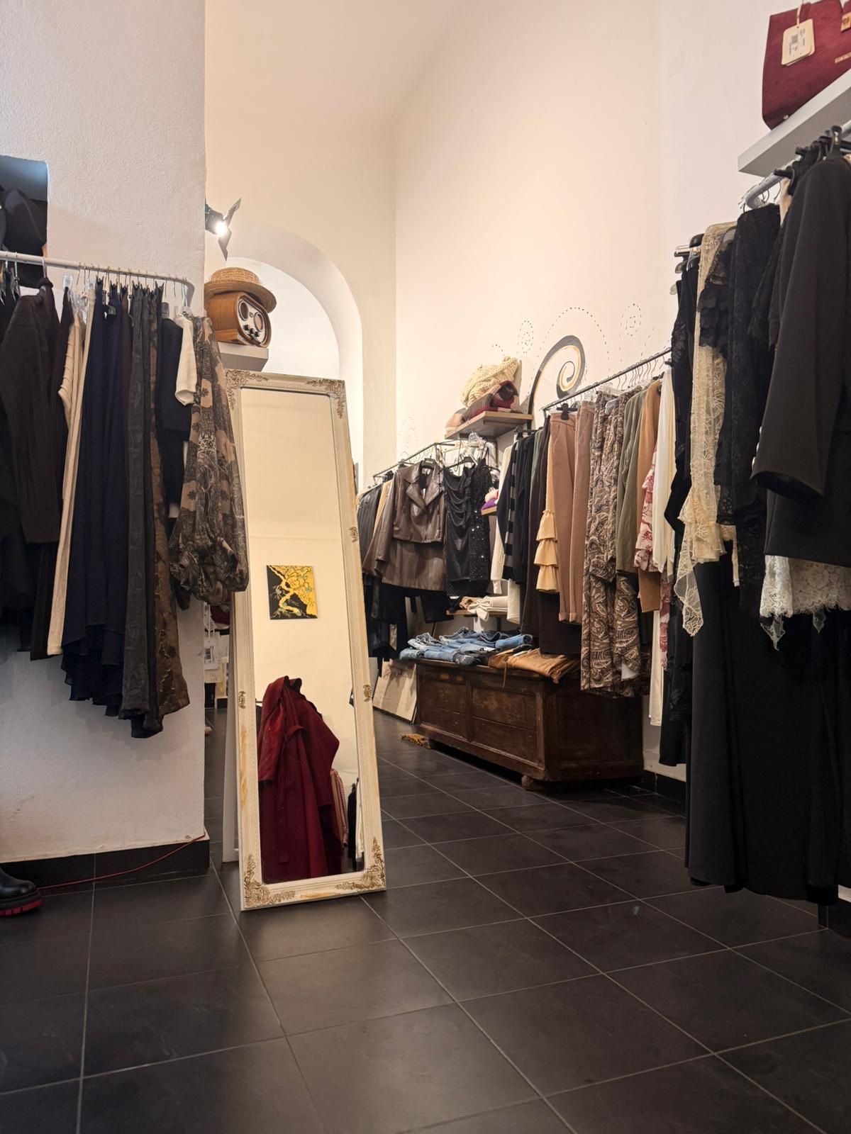 Interno di un negozio di abbigliamento con appendiabiti, uno specchio a figura intera e pavimento in piastrelle scure.