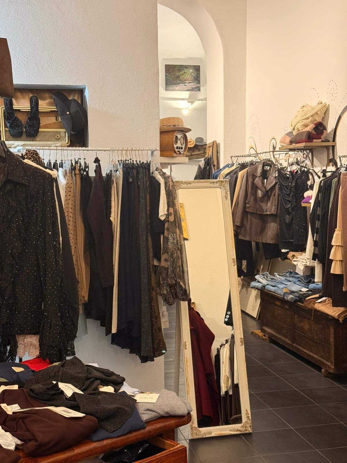 Interno di un negozio di abbigliamento, appendiabiti, specchio, cassapanca in legno, pavimenti scuri, pareti neutre, porta ad arco sullo sfondo.