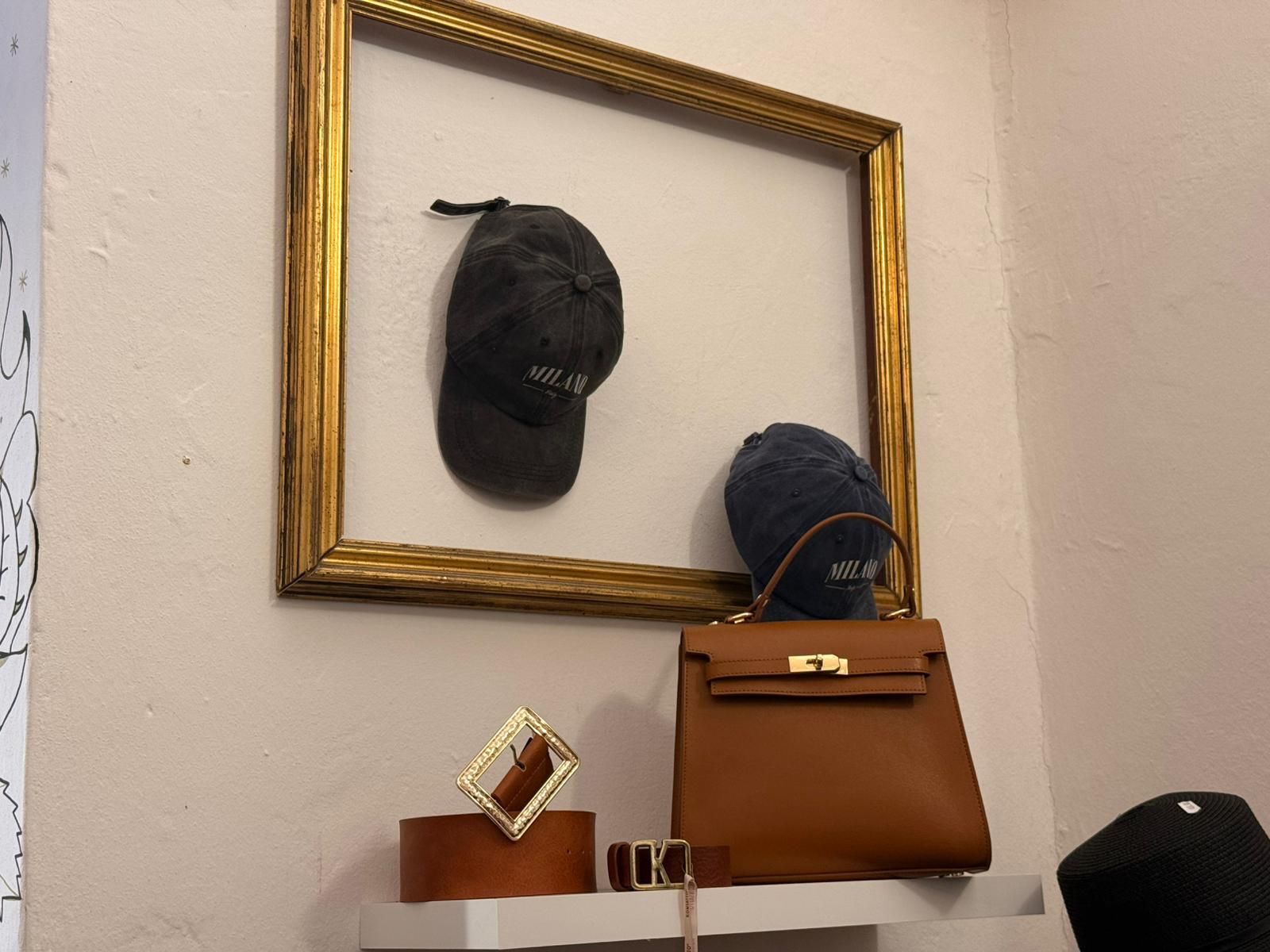 Esposizione di cappelli, una borsa e una scatola su uno scaffale all'interno di un'apertura con cornice dorata. Parete bianca.