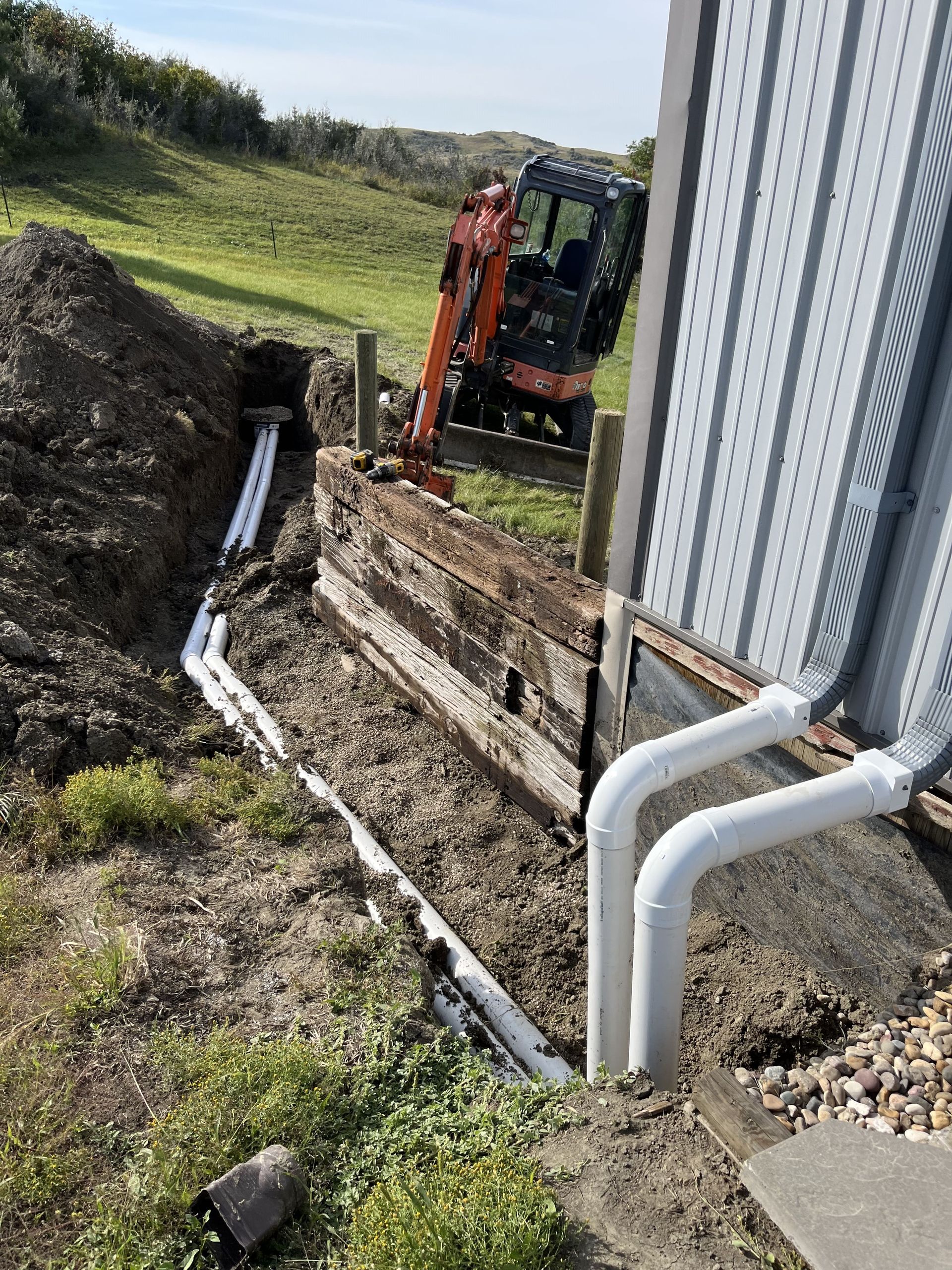 Construction site with a mini excavator, drainage pipes being installed near a building.