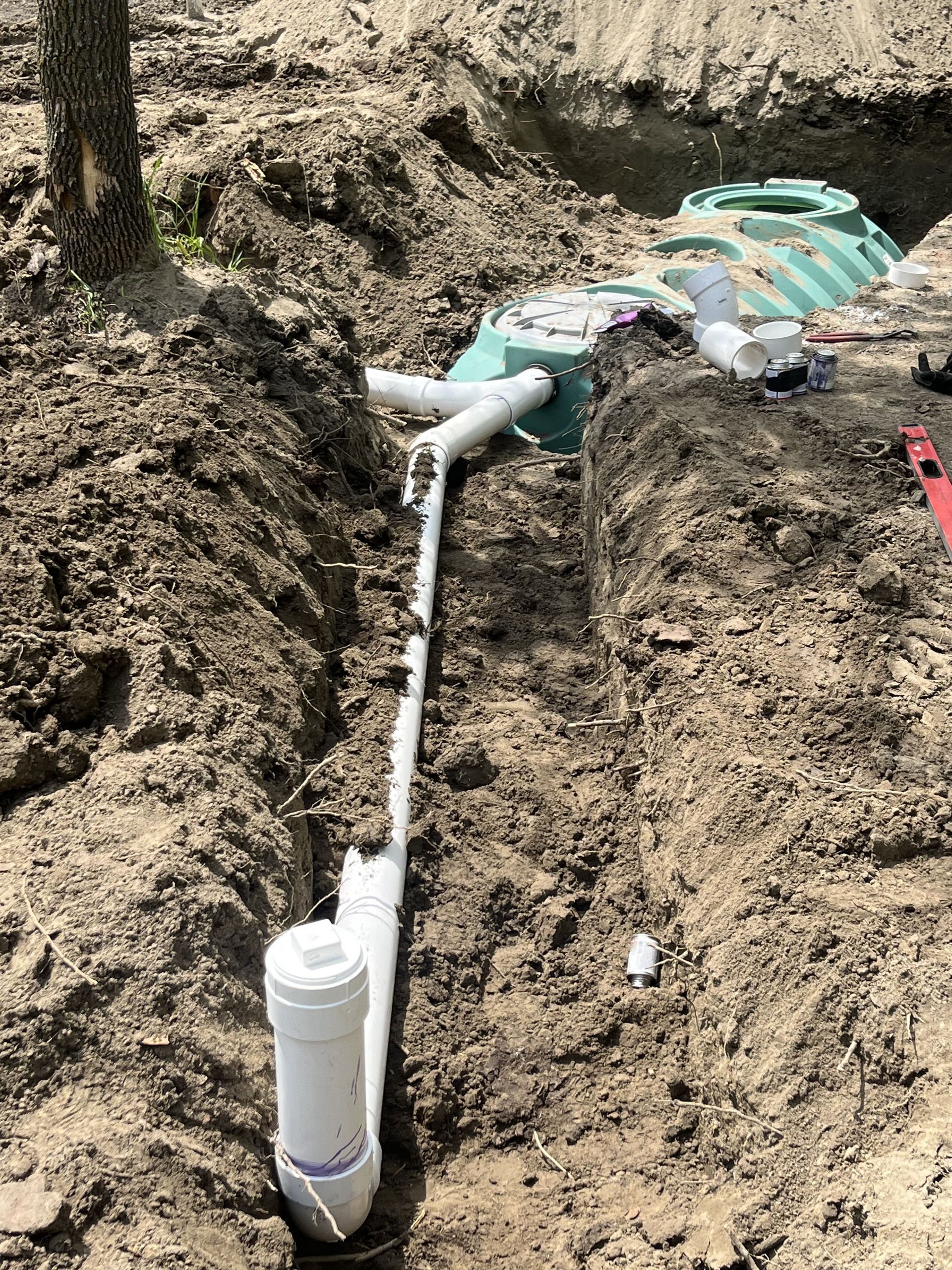 Trench dug with white pipes, fittings, and a green septic tank lid in dirt.