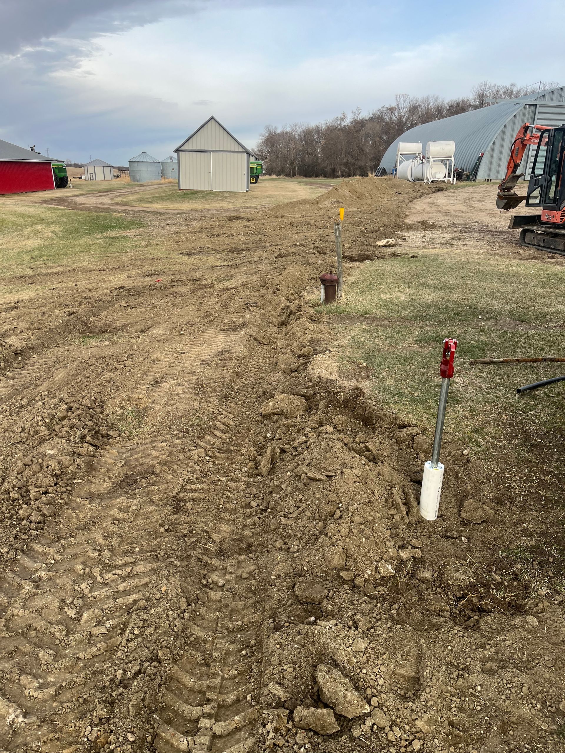 Trenched dirt path on a farm with a shovel, building, and an excavator. Overcast sky.