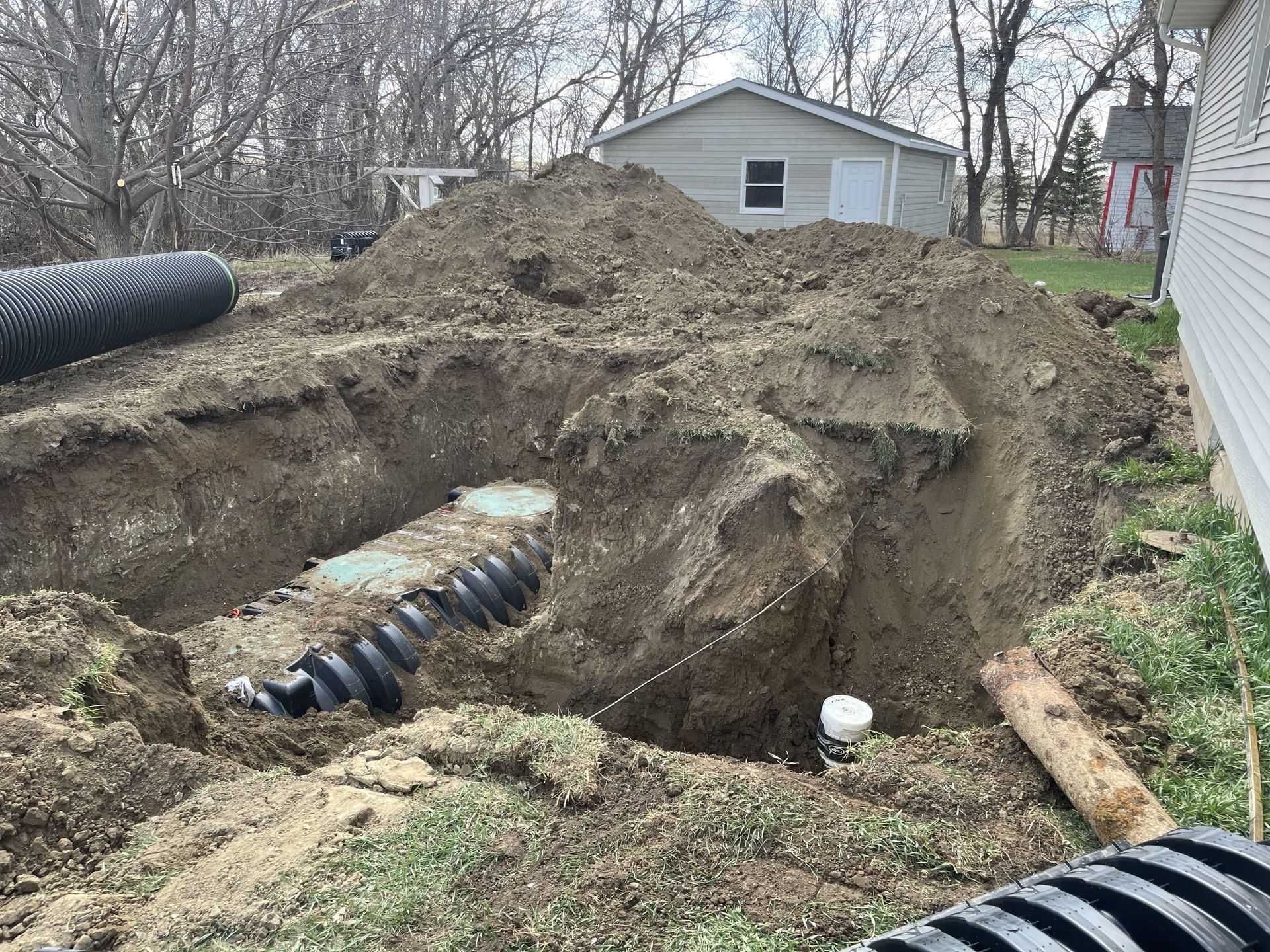Excavation site with black drainage pipes; dirt pile, white house and building in the background.