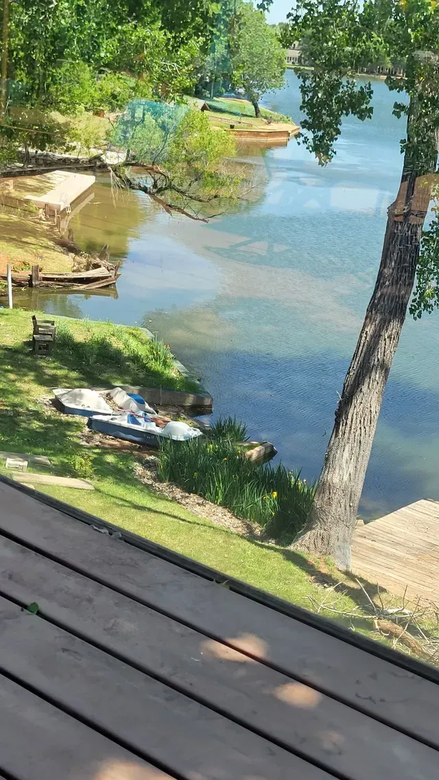 Shaded lakeside view with a docked boat, clear blue water, trees, and a wooden deck in the foreground