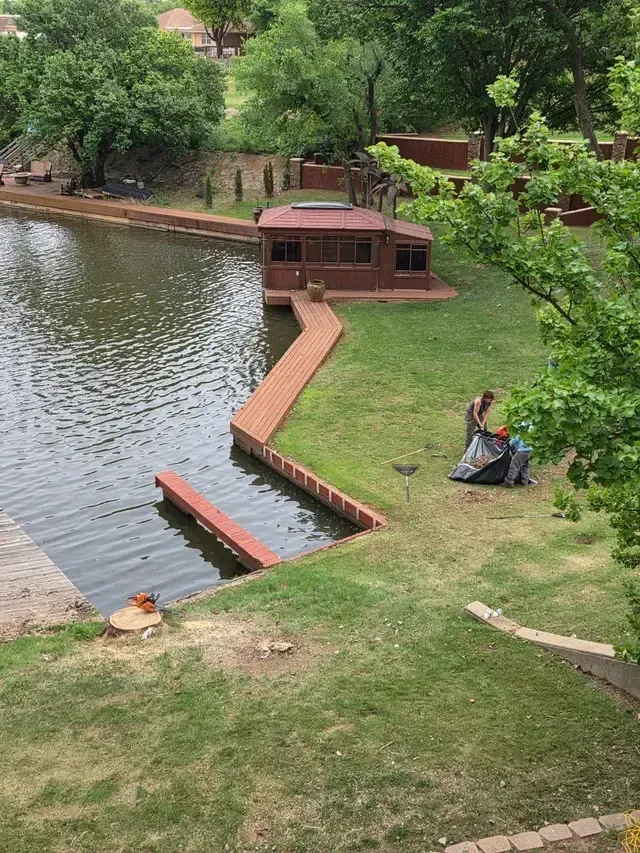 Park lake with dock and small boat; two people sit on the grassy bank nearby.