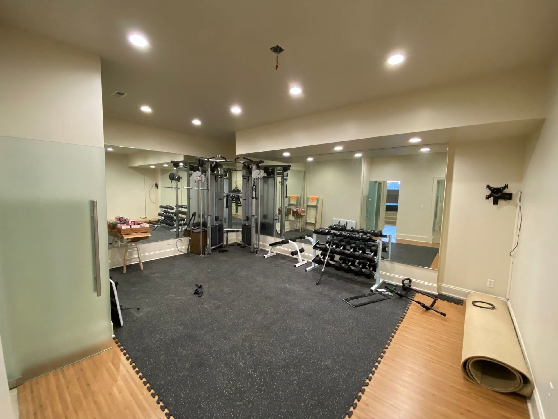 Empty gym room with exercise machines, mirrors, and dark carpeted flooring under recessed ceiling lights