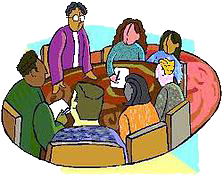 קבוצת אנשים יושבת סביב שולחן עגול, אחד עומד, שקוע בדיון.