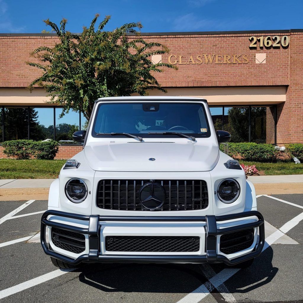 A white mercedes benz g63 amg is parked in a parking lot in front of a brick building.