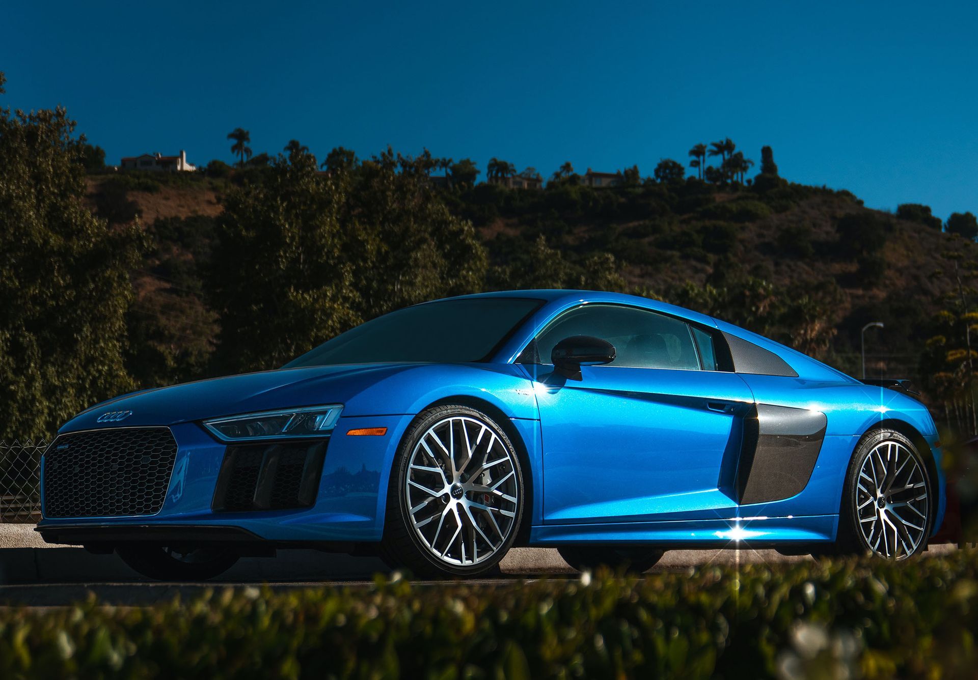 Blue Audi R8 sports car parked on a road, with dark wheels, a green hedge, and a blue sky in the background.