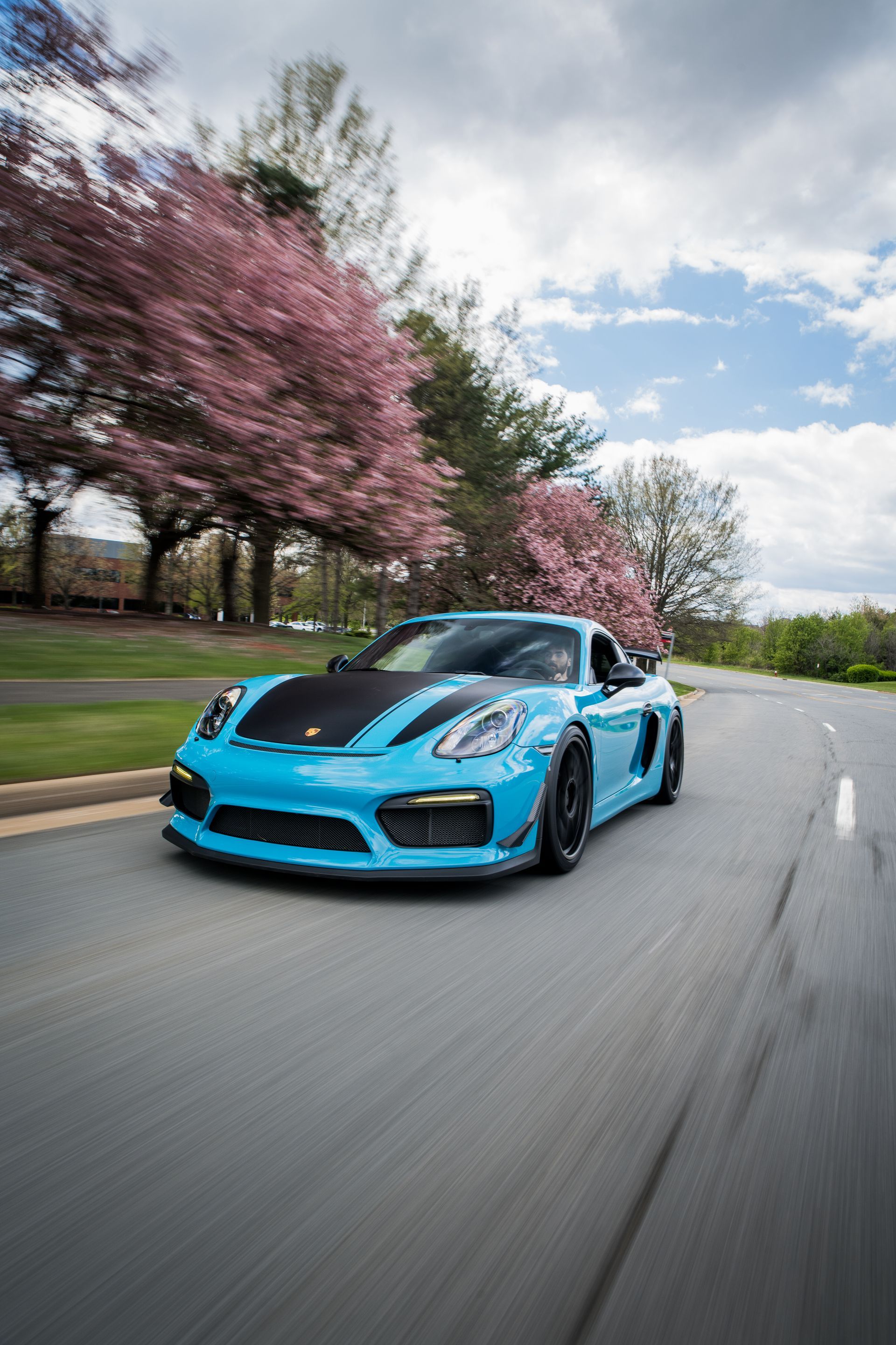 A blue porsche boxter gt4 is driving down a road with cherry blossom trees in the background.