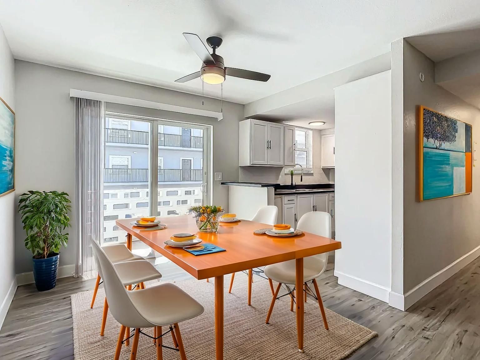 Dining room with orange table, six white chairs, and open kitchen. Large windows and a ceiling fan.