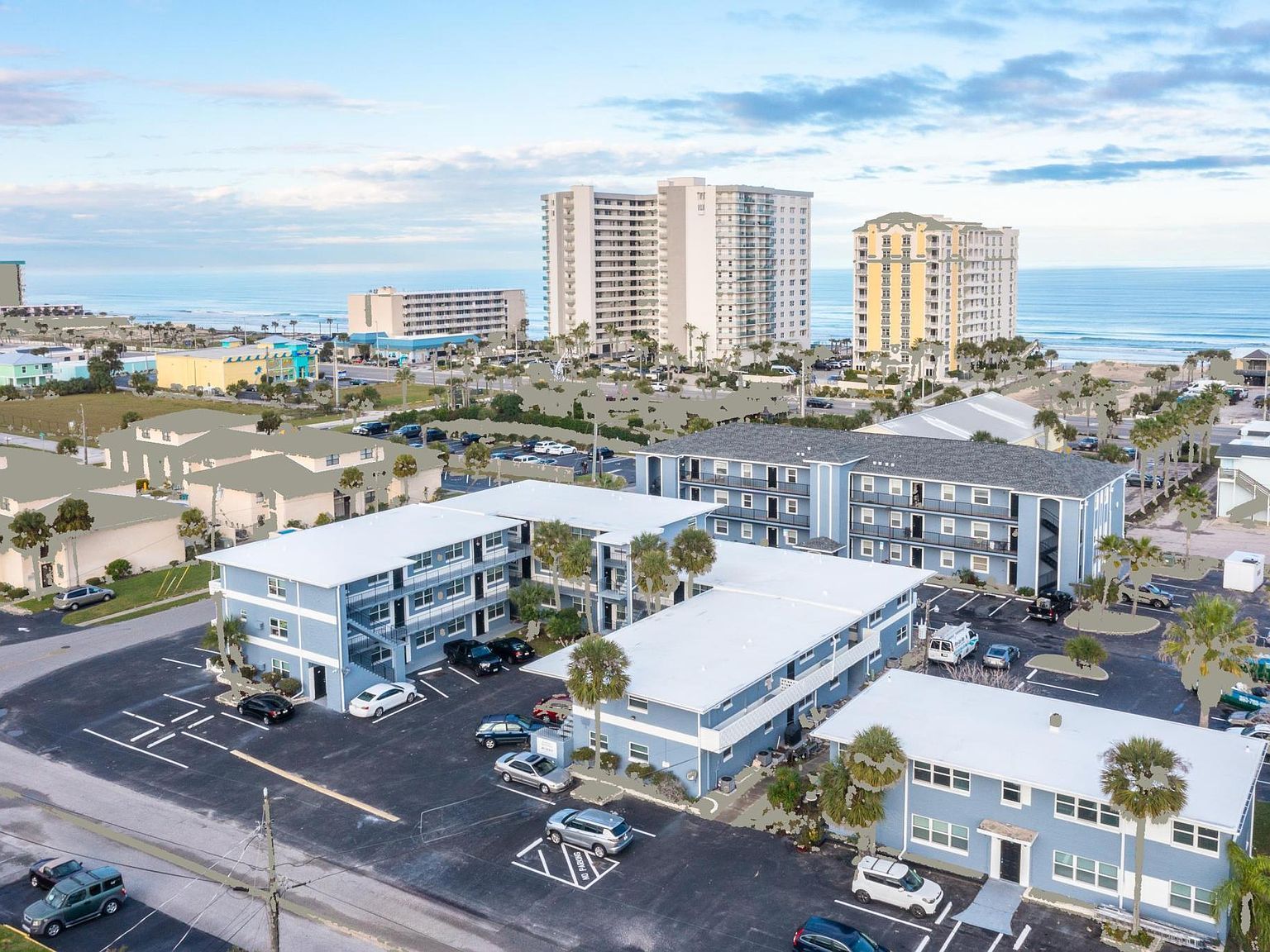 Aerial view of beachside apartments and condos. Blue and white buildings with ocean and sky in background.