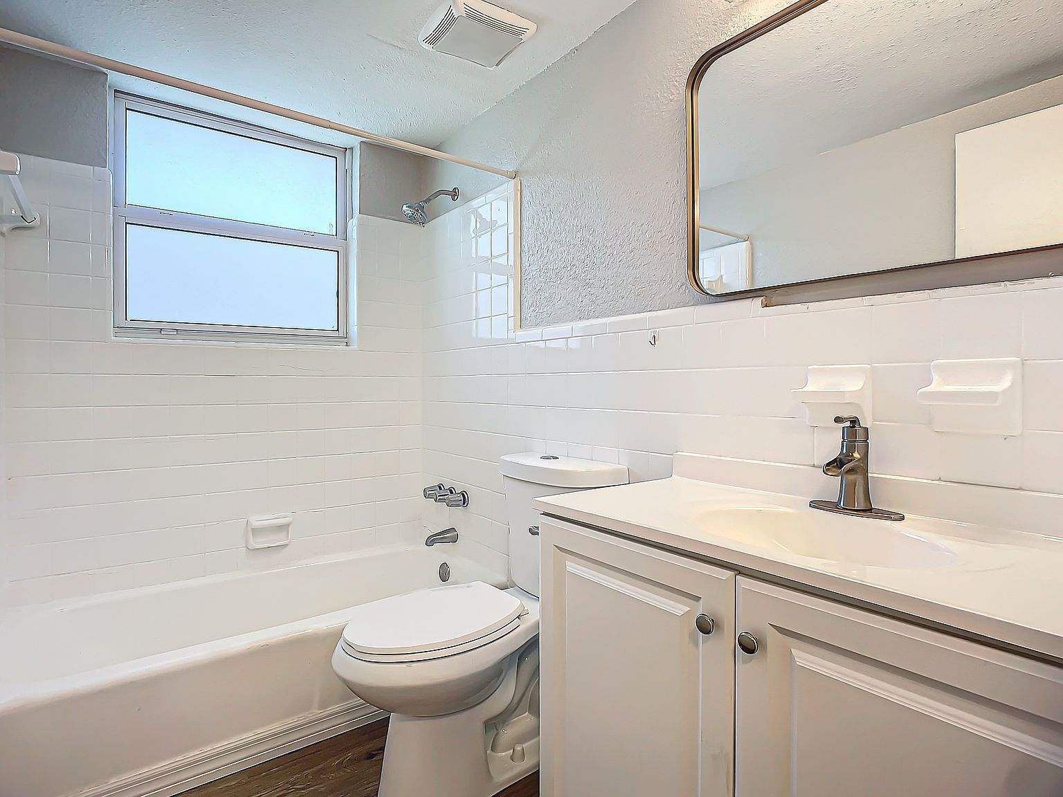 White bathroom with tub, toilet, vanity, and window. Mirror reflects the room.