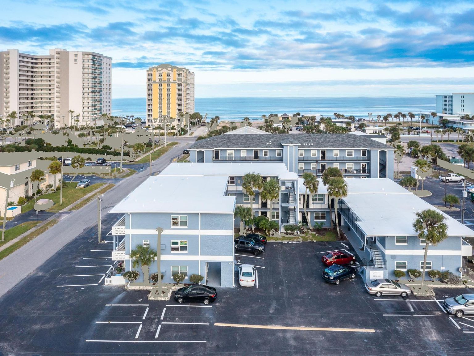 Aerial view of a light blue apartment complex with parking, near a beach with high-rise buildings in the background.