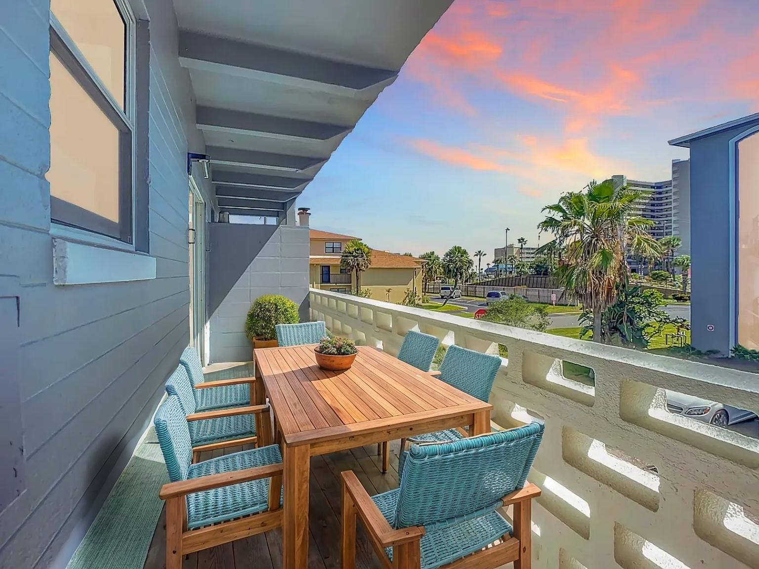 Balcony with wooden table, blue chairs, ocean view, and sunset sky.