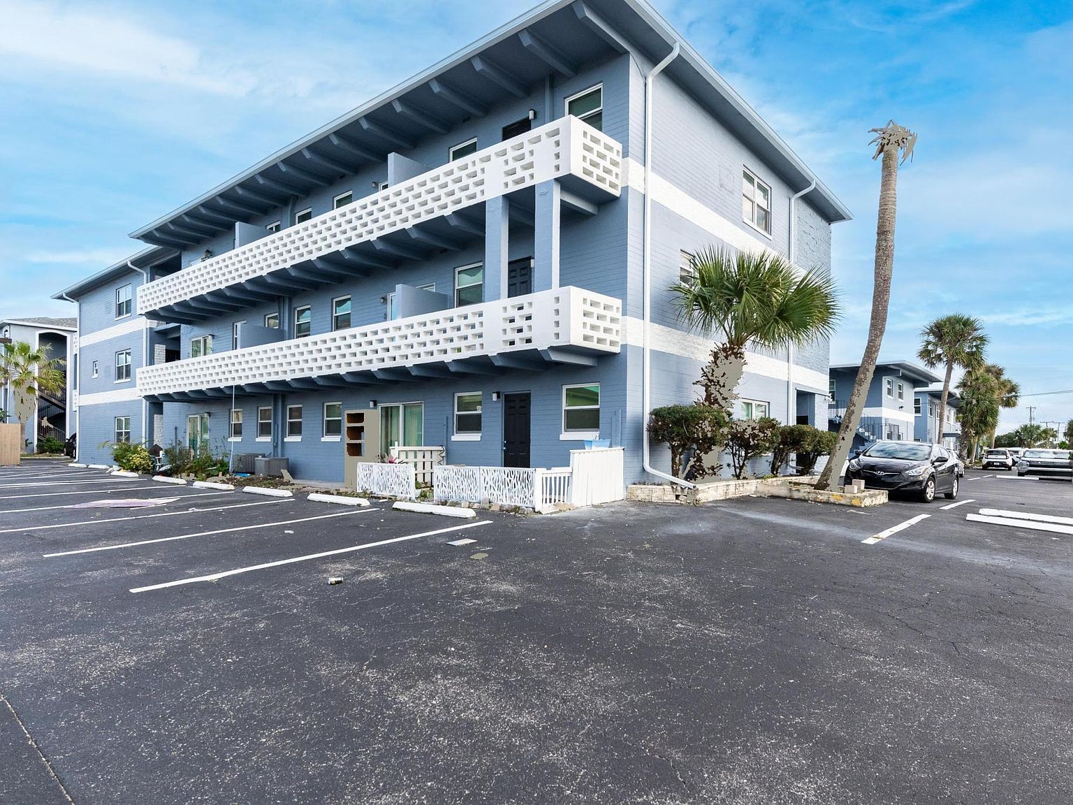 Blue multi-story condo building with parking lot and palm trees.