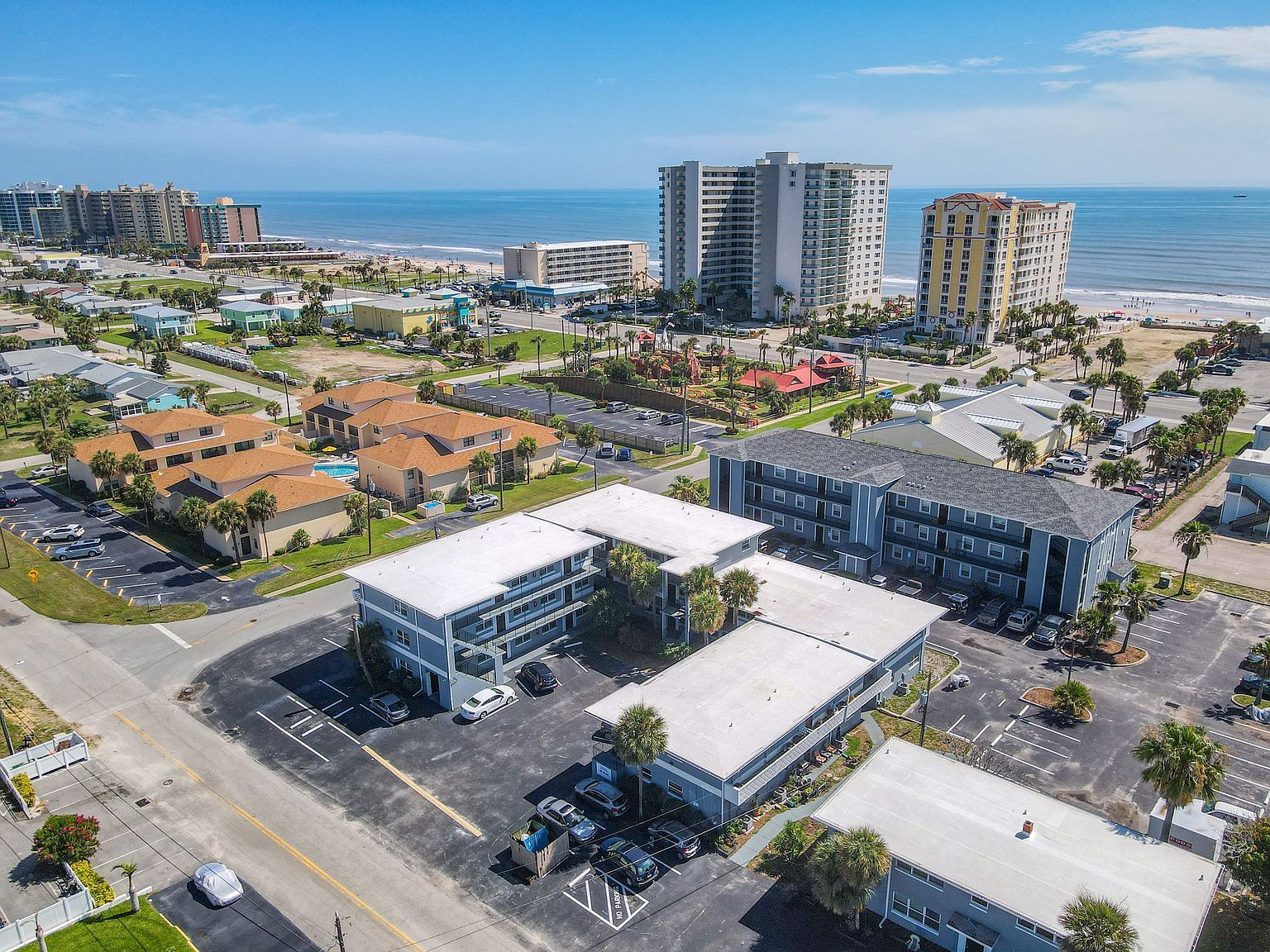 Aerial view of a coastal hotel complex with ocean and buildings in the background. Bright sunny day, palm trees.