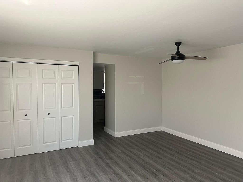 A mostly empty room with white closet doors, gray walls, gray flooring, and a ceiling fan.