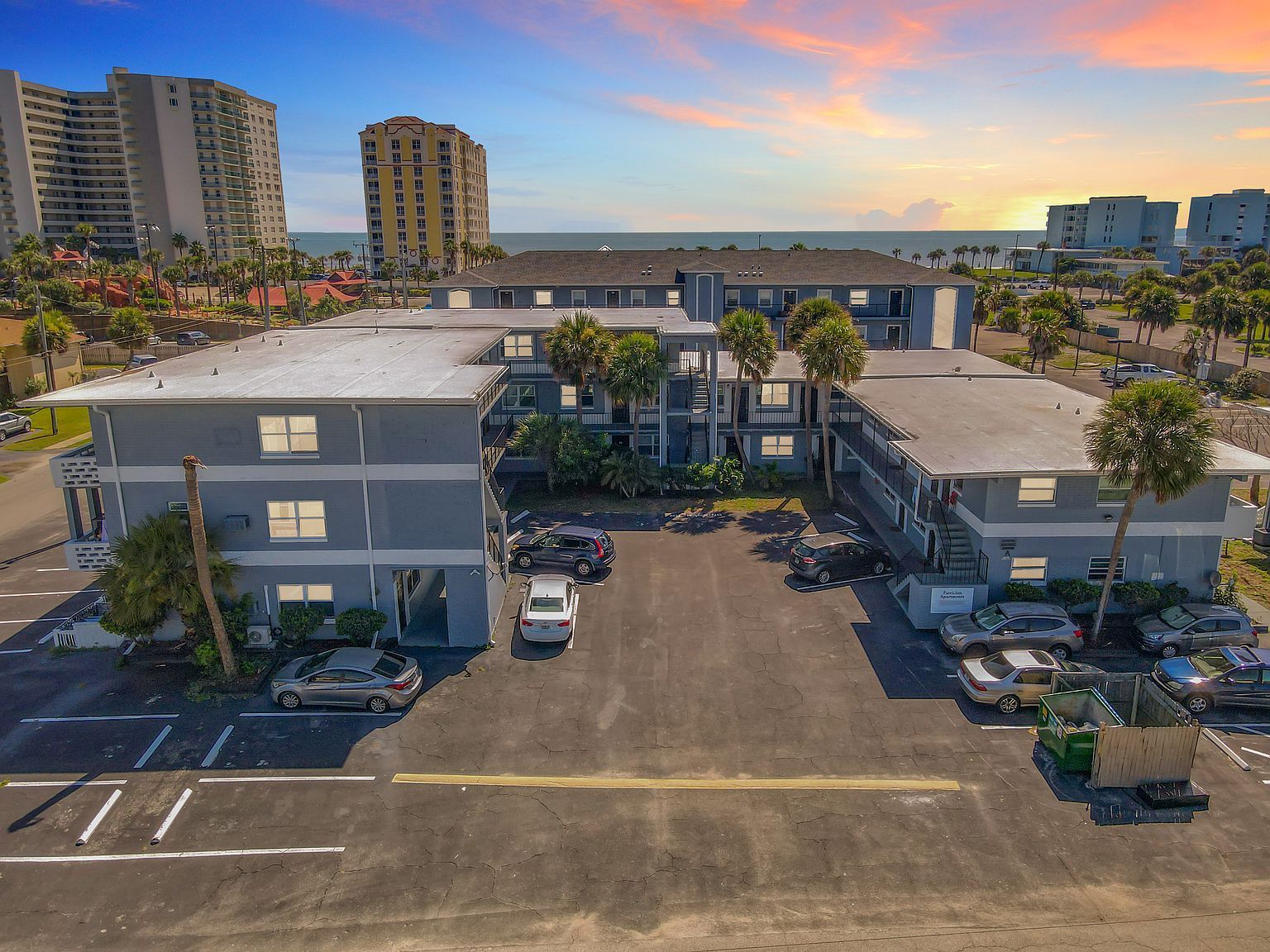 Exterior aerial view of a gray two-story apartment complex with parking, near a beach at sunset.