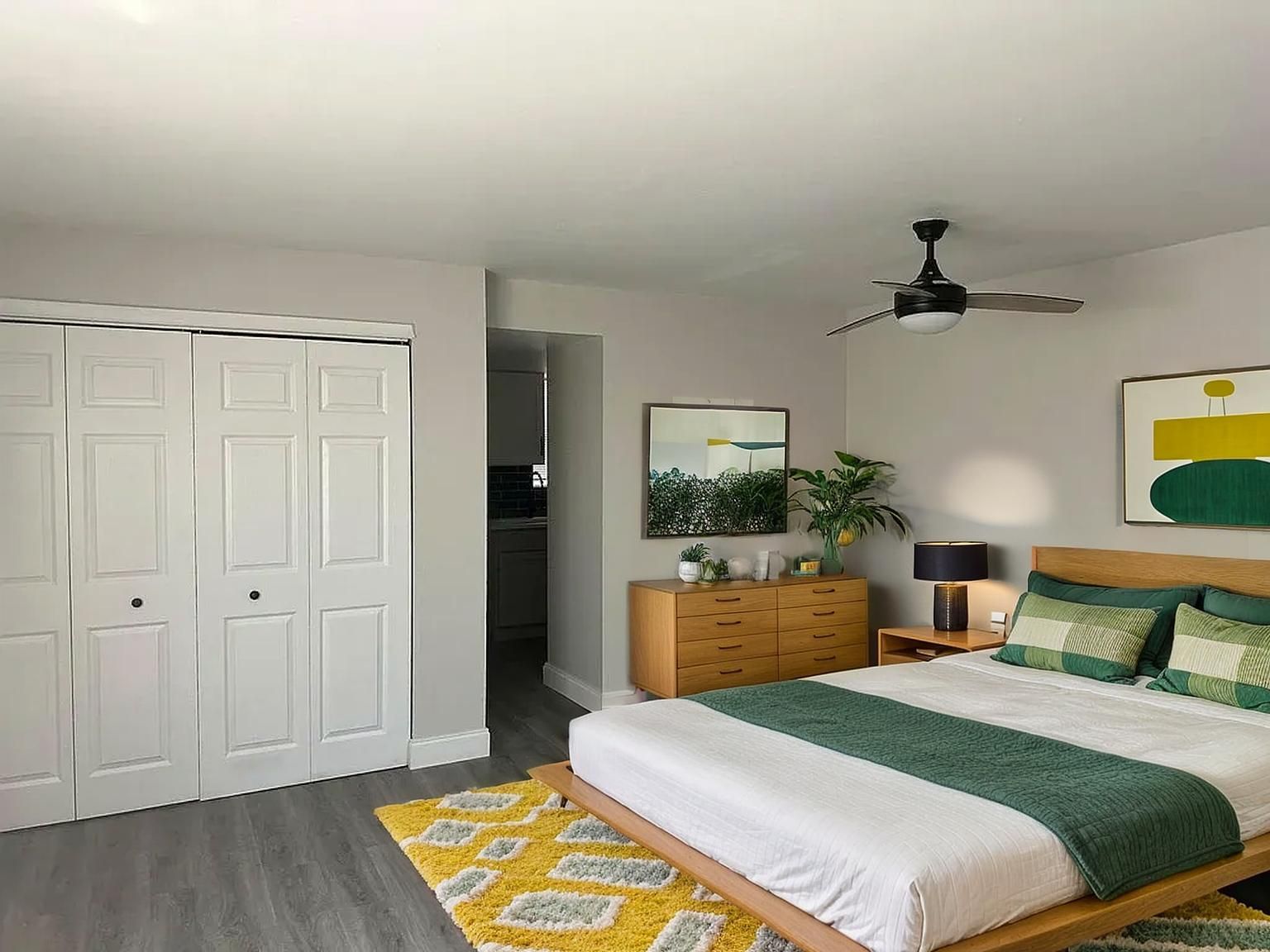 Bedroom with wooden bed, dresser, art, and yellow rug. White closet doors. Gray walls.