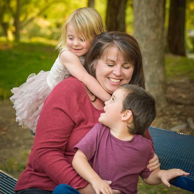 Woman smiles, embraced by a child with arms around her neck and another child in her lap, outdoors.
