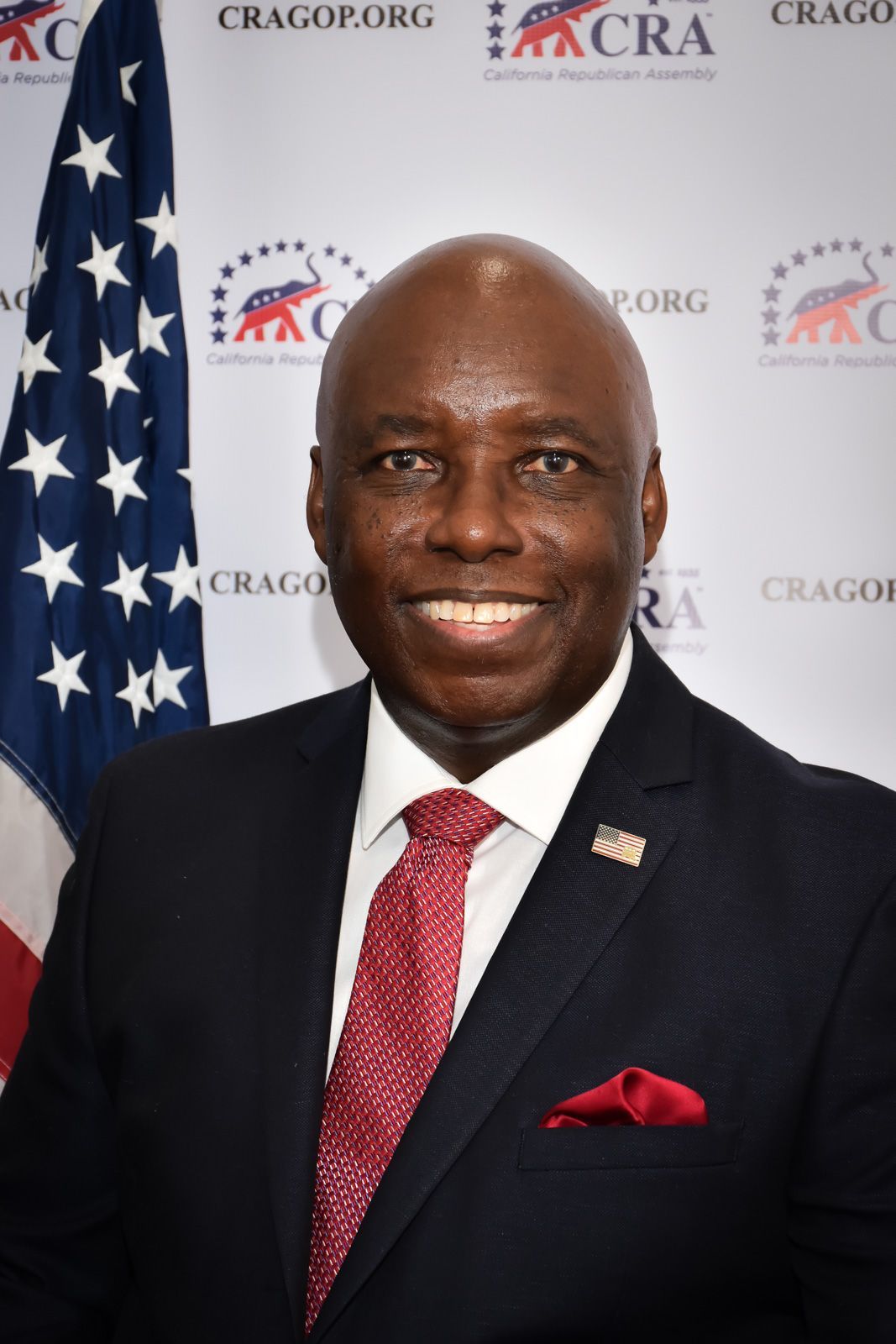 Man in a suit smiles, standing in front of an American flag and a Republican Party logo.