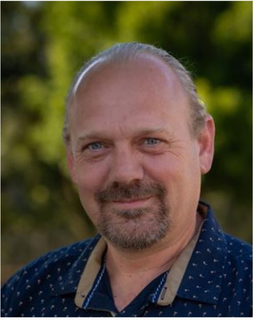 Man with light skin and a beard, smiling, wearing a dark blue shirt, outdoors.