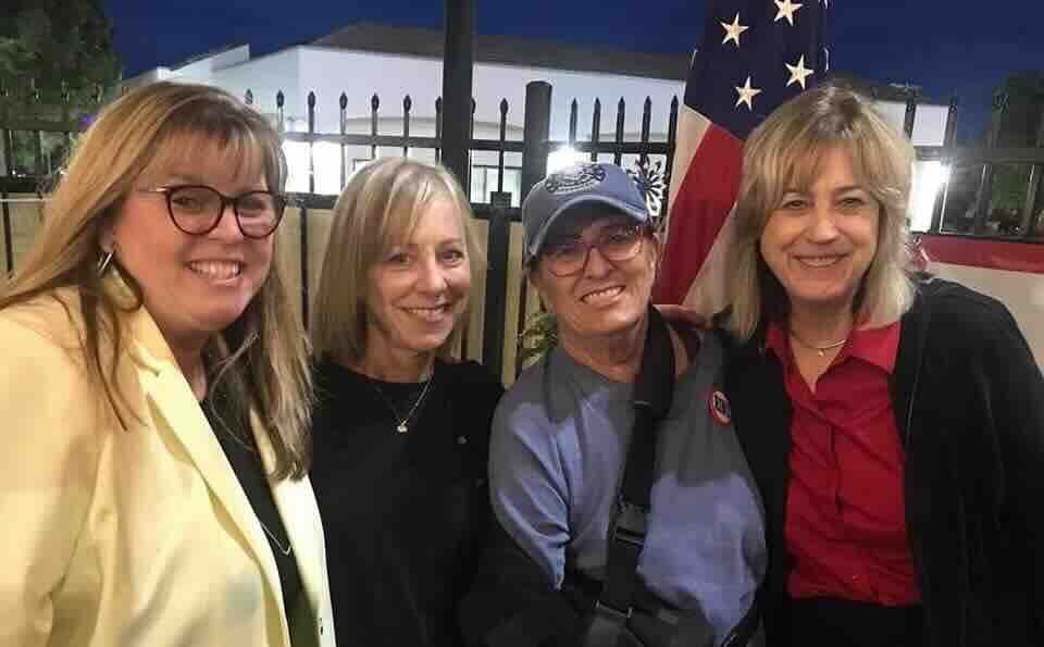 Four women smiling, posing for a photo. American flag in the background. Evening setting.