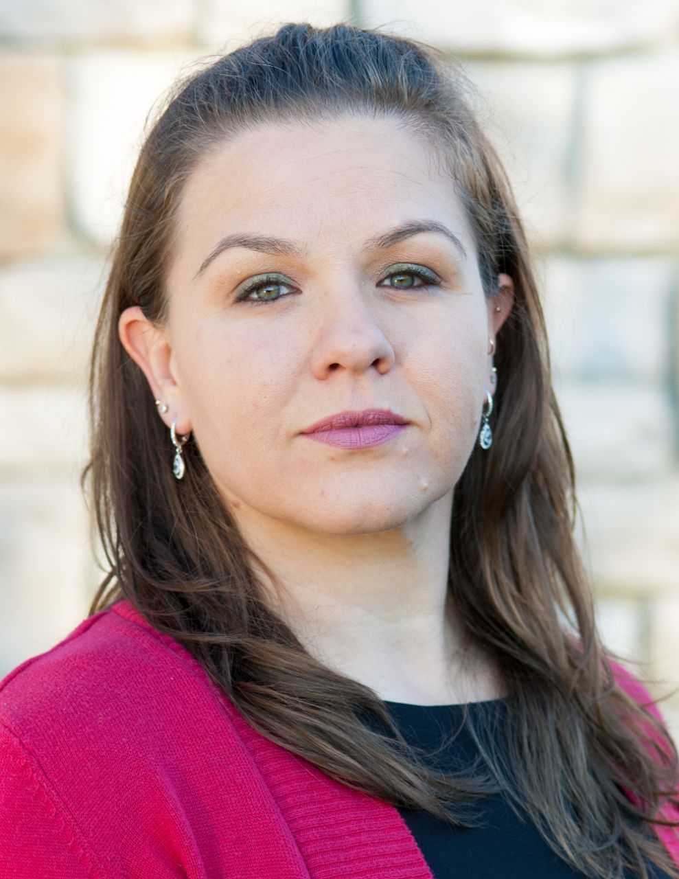 Woman in red jacket, looking at the camera, against a stone wall background.
