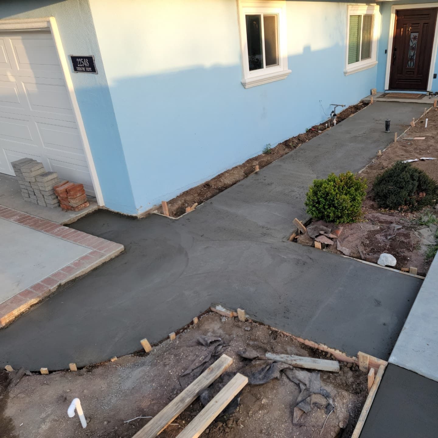 Freshly poured concrete walkway in front of a blue house, framed by wooden supports and dirt.
