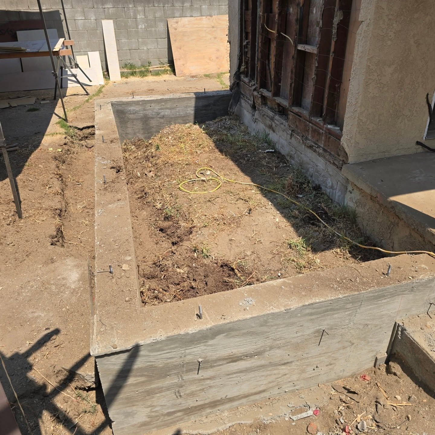 Concrete planter box with dirt and debris, near a building's partially exposed wood frame, outside.
