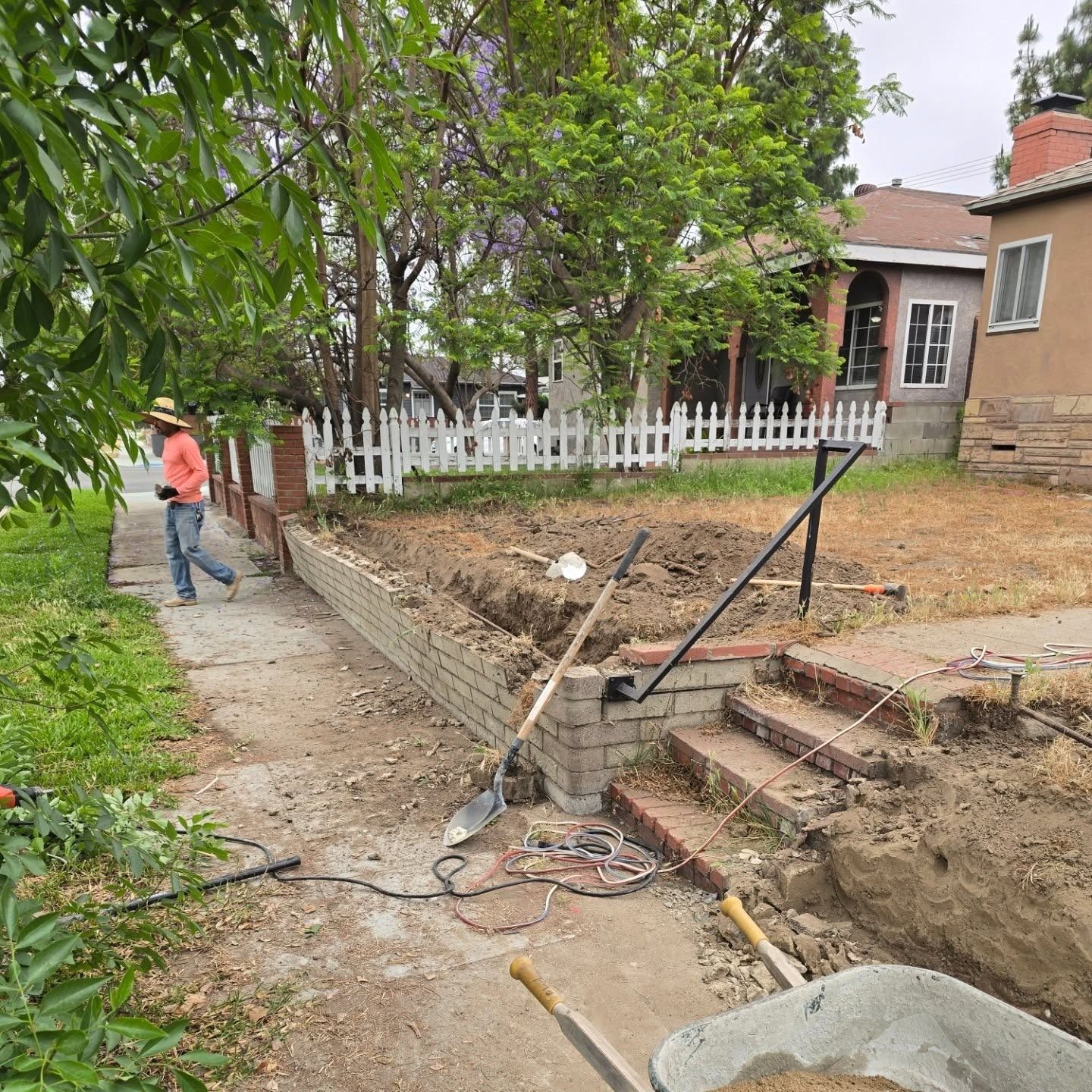 Person working on brick wall in a yard with shovel, wheelbarrow, and steps, fence in background.