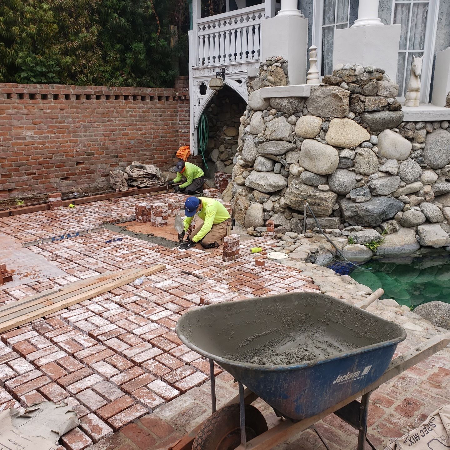 Construction workers laying brick patio next to a stone water feature. A wheelbarrow is in the foreground.