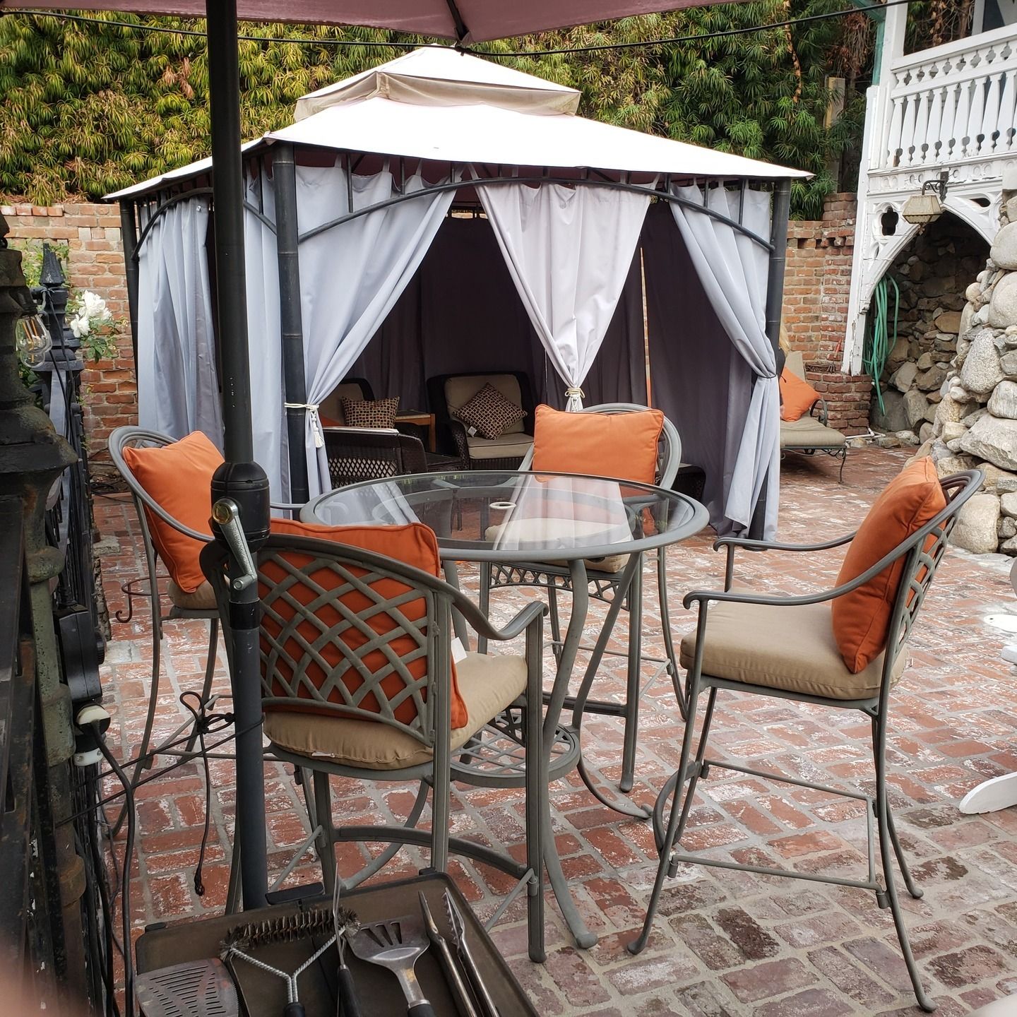Patio with glass table, chairs, orange cushions, and a gazebo with curtains on a brick patio.