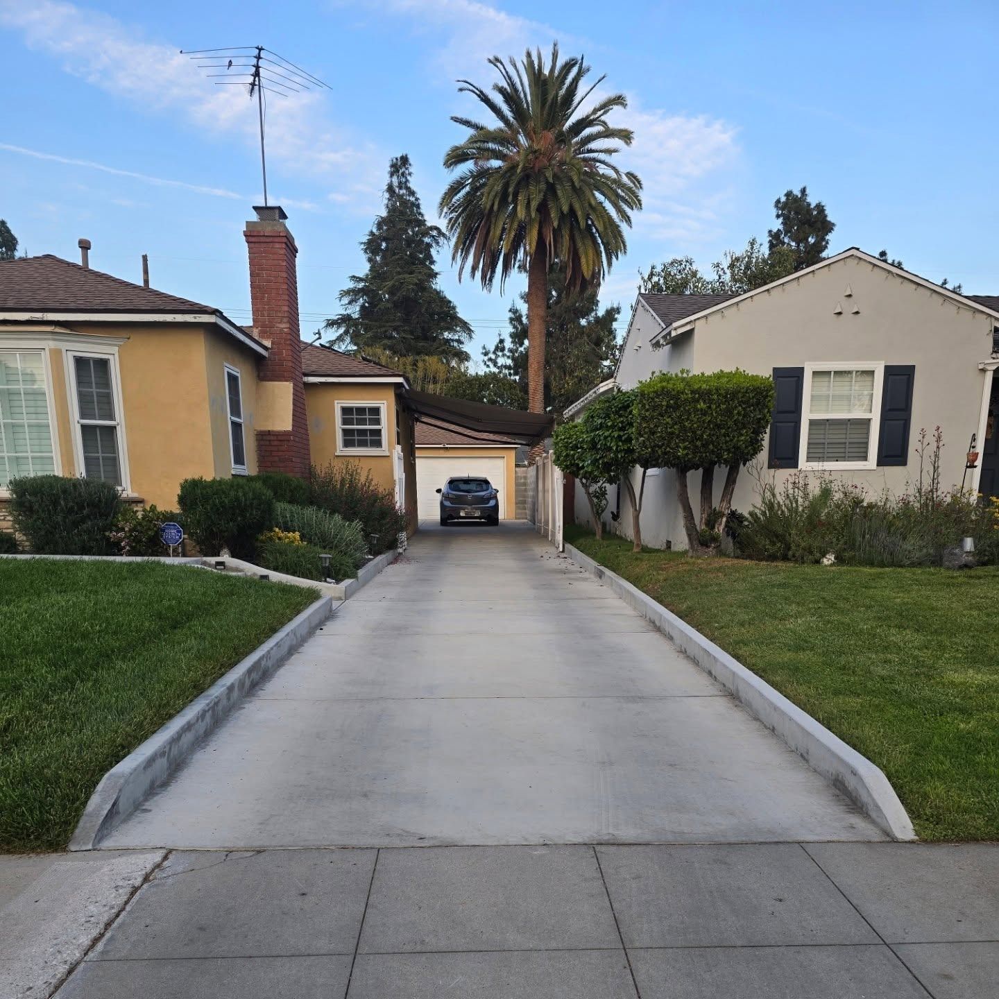 Driveway leading to a garage with a car, between two houses, one yellow and one white, palm tree in the background.