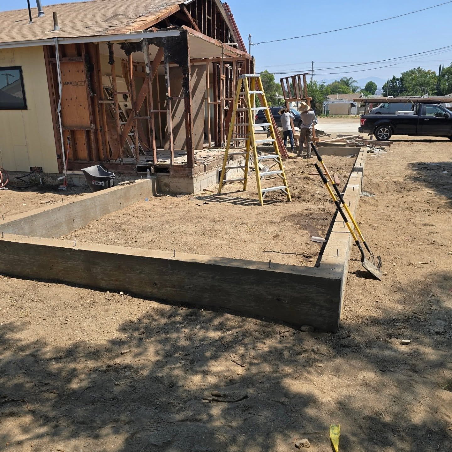 Construction site: concrete foundation in foreground, wood frame of building under construction, yellow ladder.