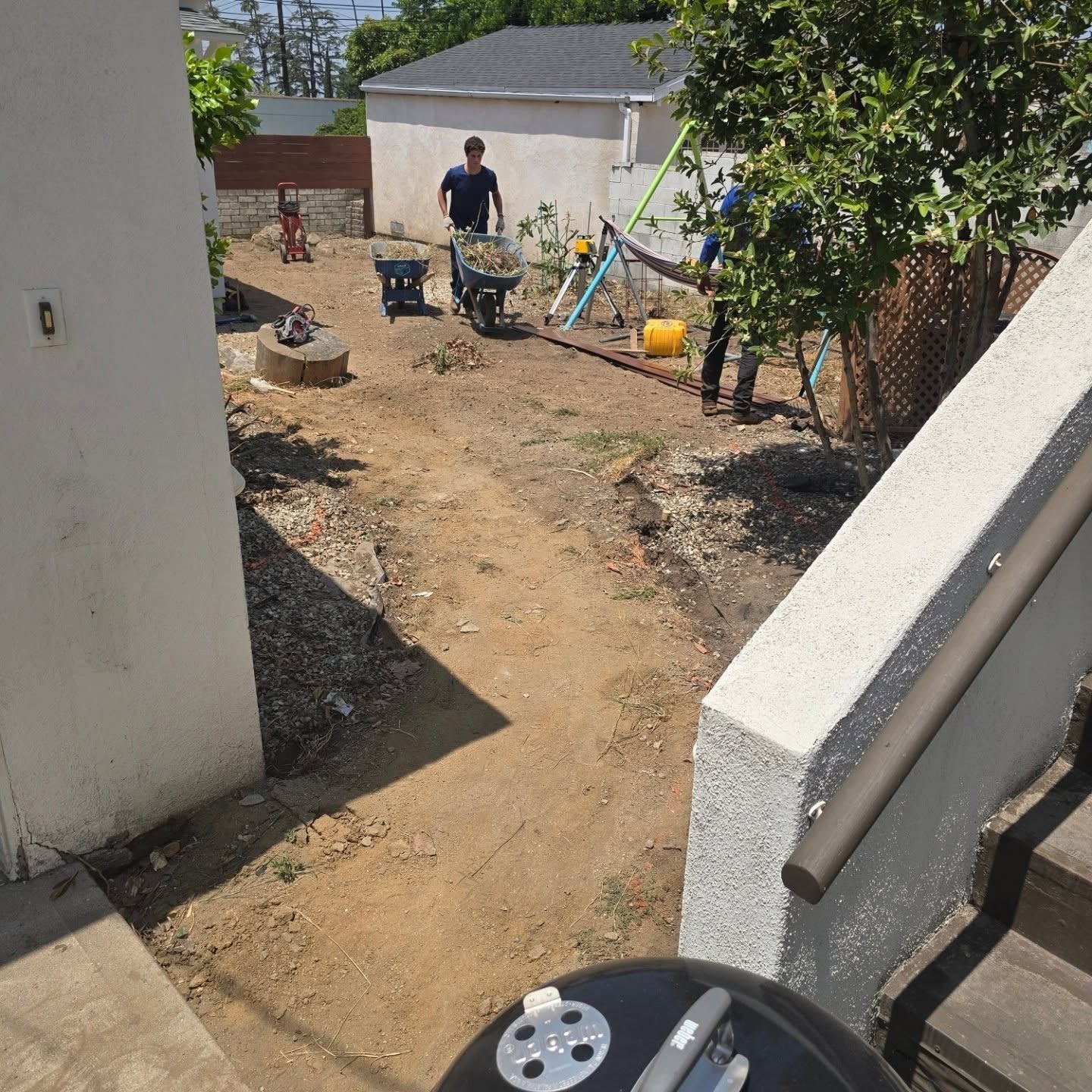 People working in a dirt backyard, using a wheelbarrow and tools near a building, on a sunny day.
