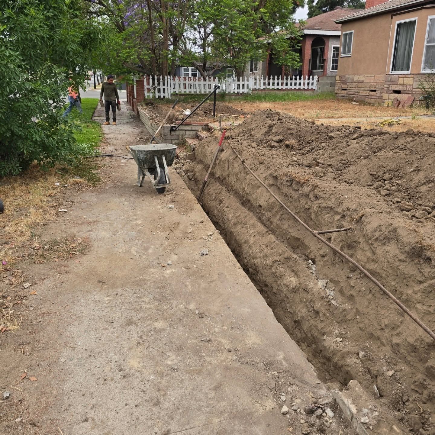 A trench dug in a dirt path, with a wheelbarrow, person walking, houses in background.