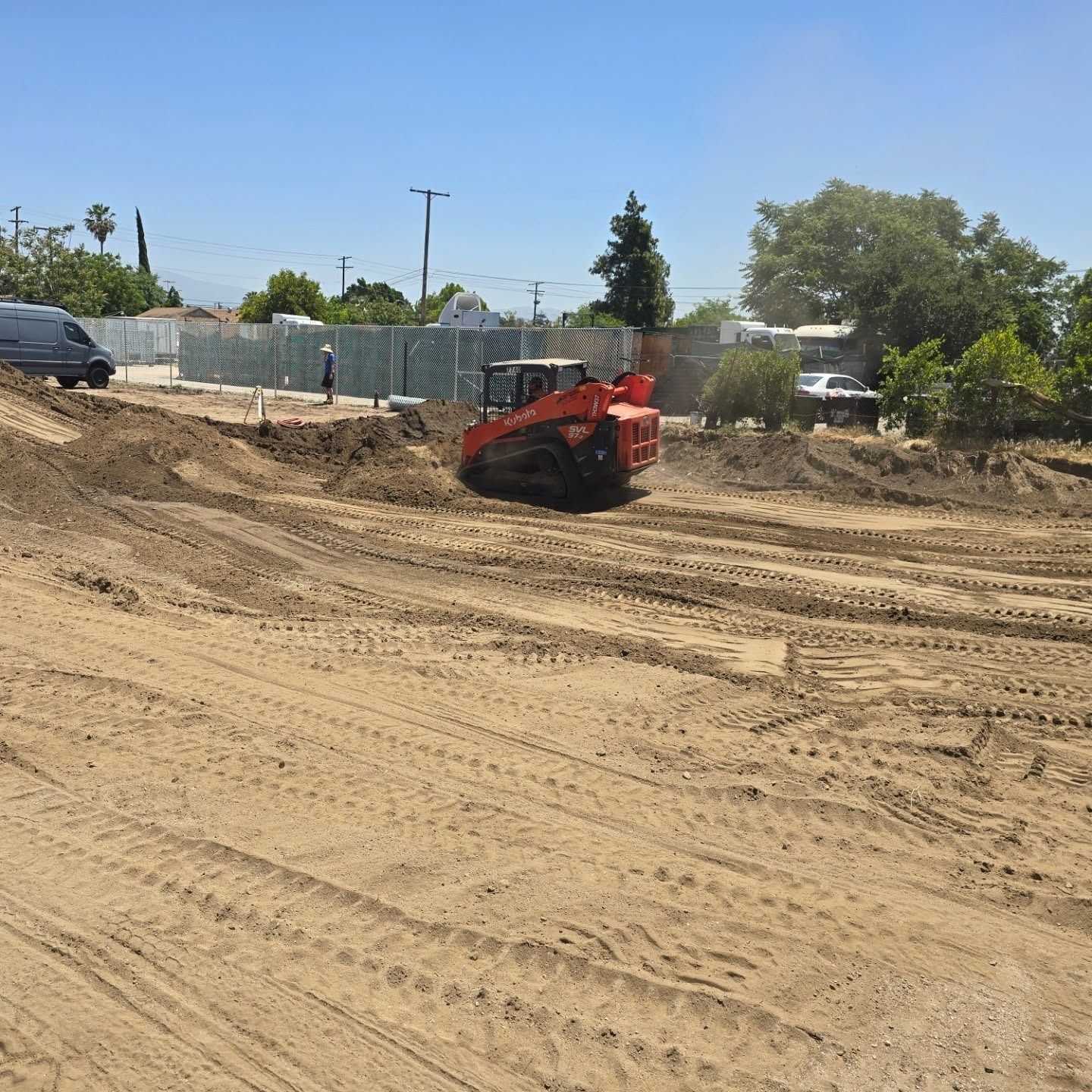 Orange skid steer grading a dirt lot under a clear blue sky.