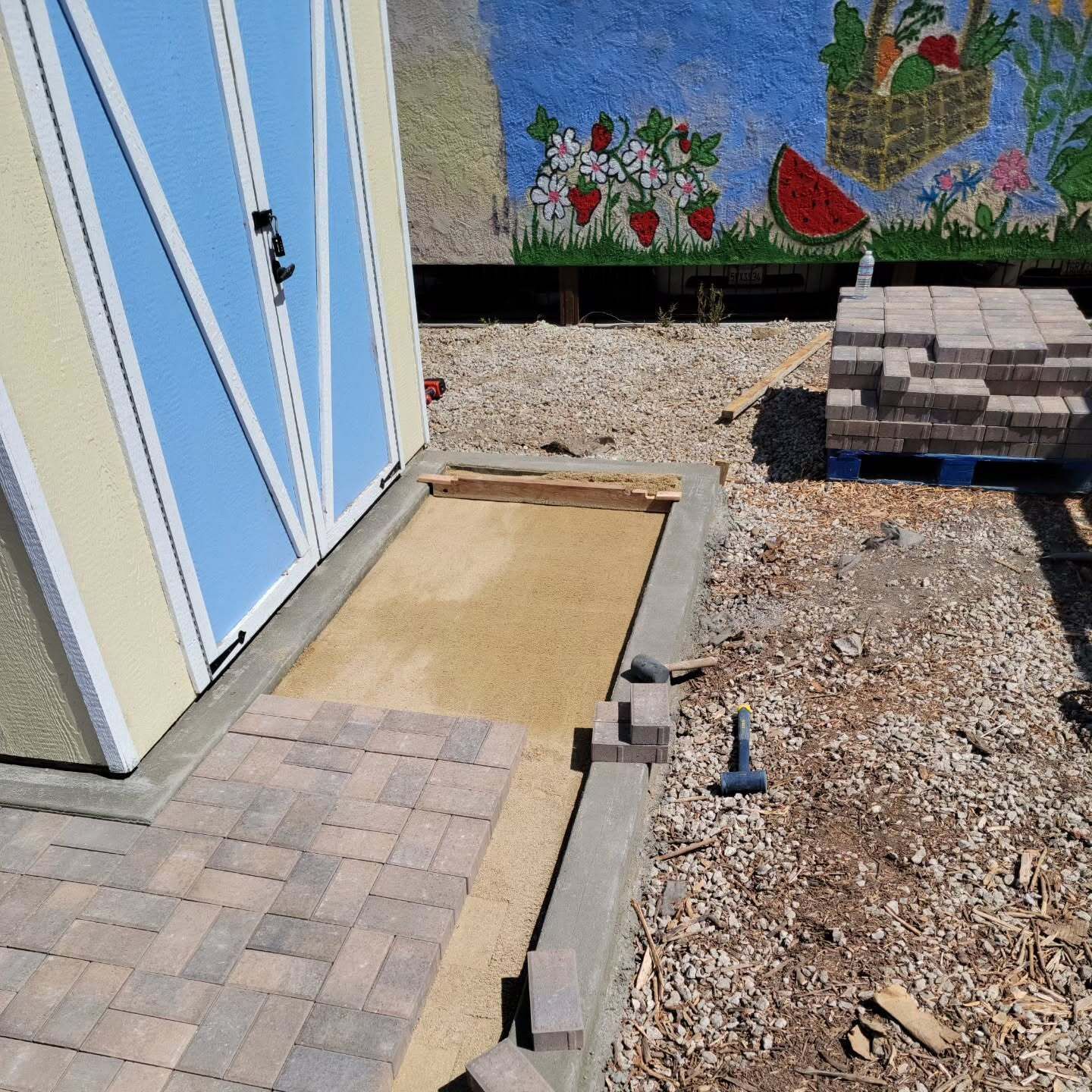 Construction site: a shed with a blue door, sand, pavers, and stacked bricks outdoors.