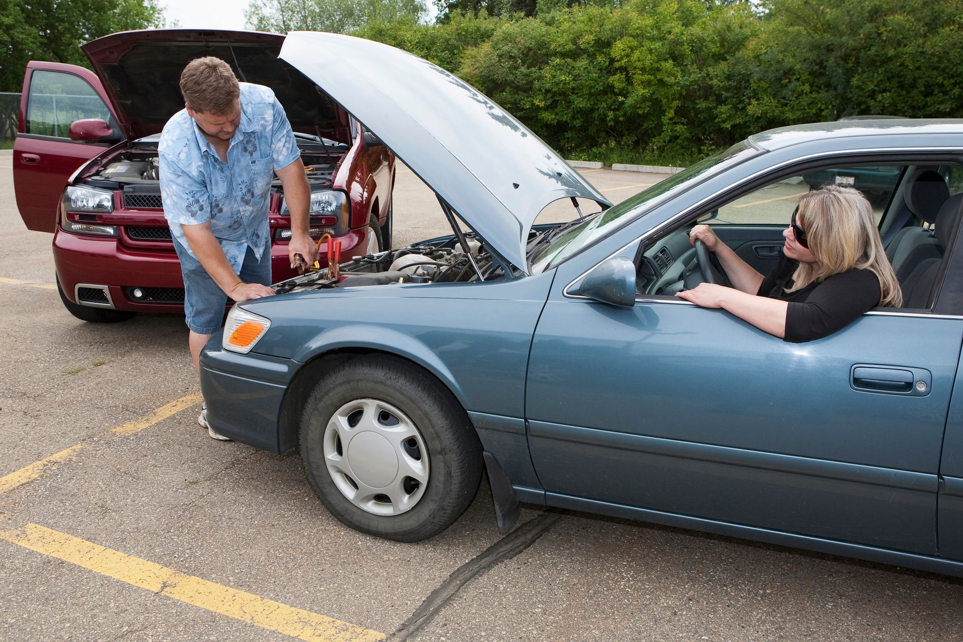 A man jumps a car's battery for a woman in the driver's seat; both cars have open hoods in a parking lot. A man jumps a car's battery for a woman in the driver's seat; both cars have open hoods in a parking lot.